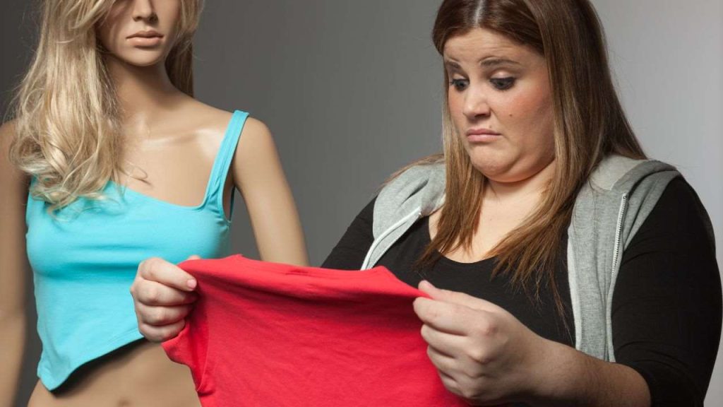 An image showing a woman looking concerned as she holds a red shirt, seemingly dissatisfied with its fit. Beside her is a mannequin dressed in a blue tank top. The scene captures the feelings of frustration when clothing sizes do not meet expectations.